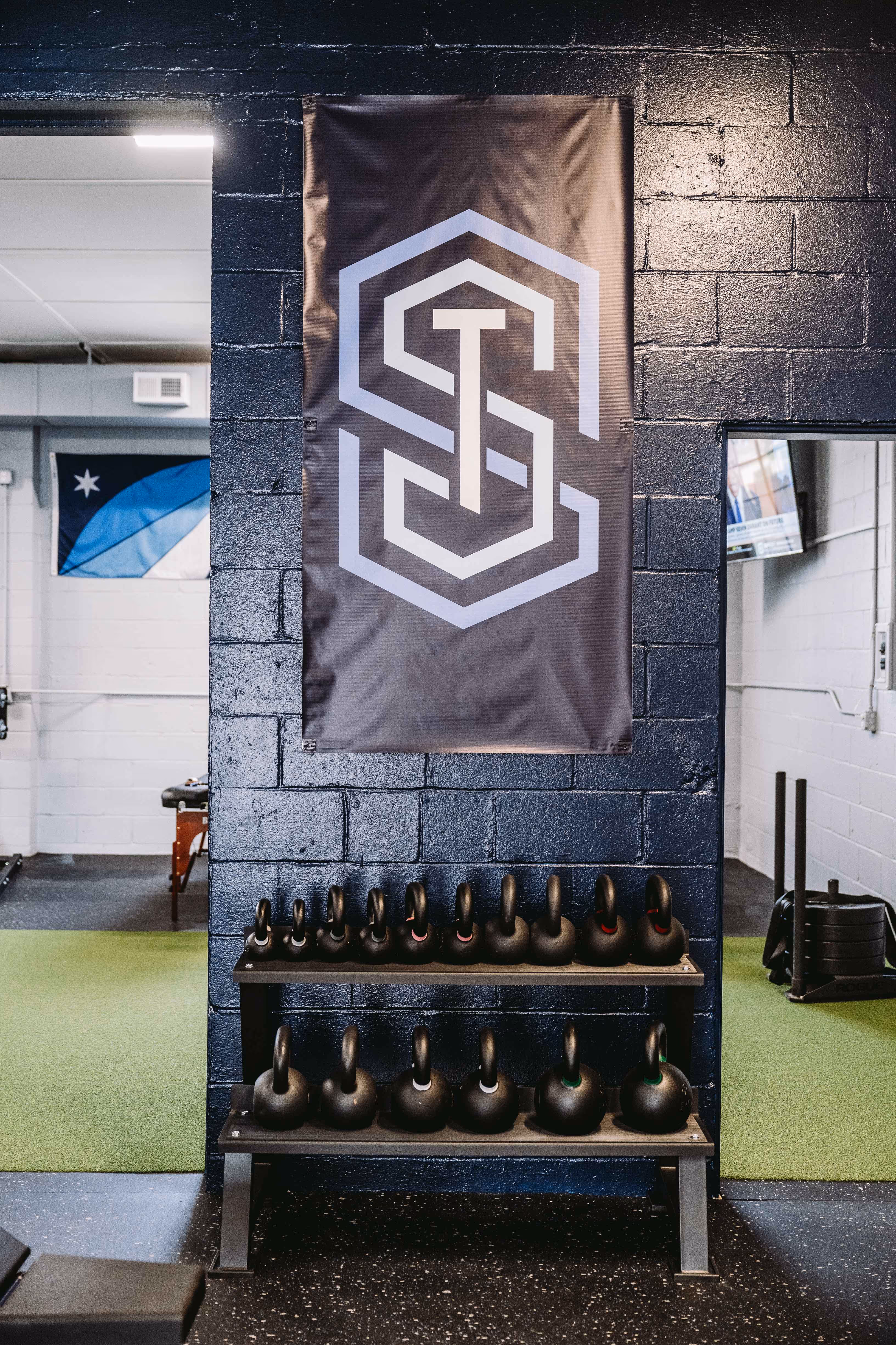 Two-tier rack holding multiple black kettlebells against a dark blue brick wall with a large logo banner above in a gym setting.