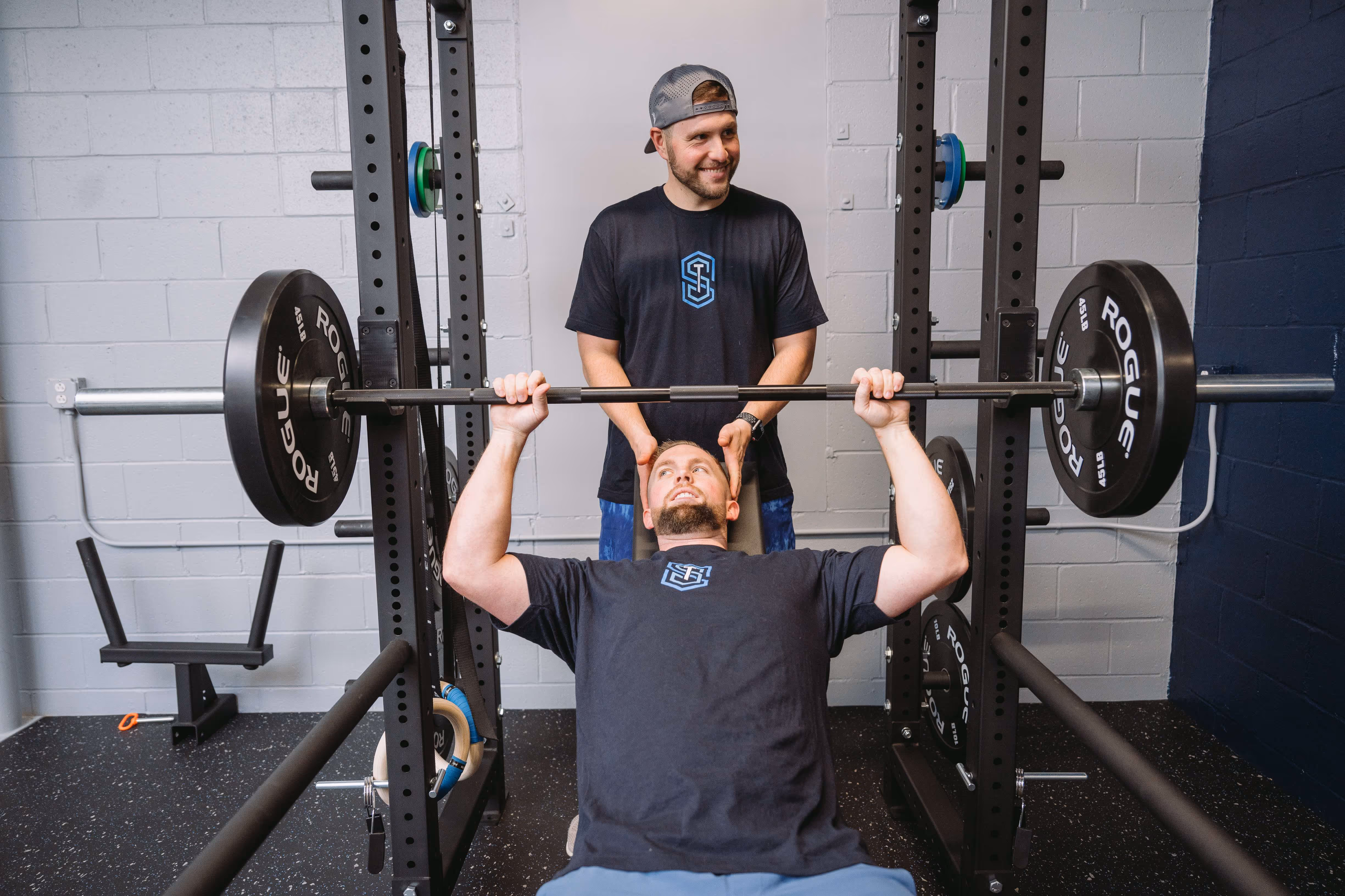 Man in black t-shirt performing bench press with barbell while another man stands behind spotting him in a gym.