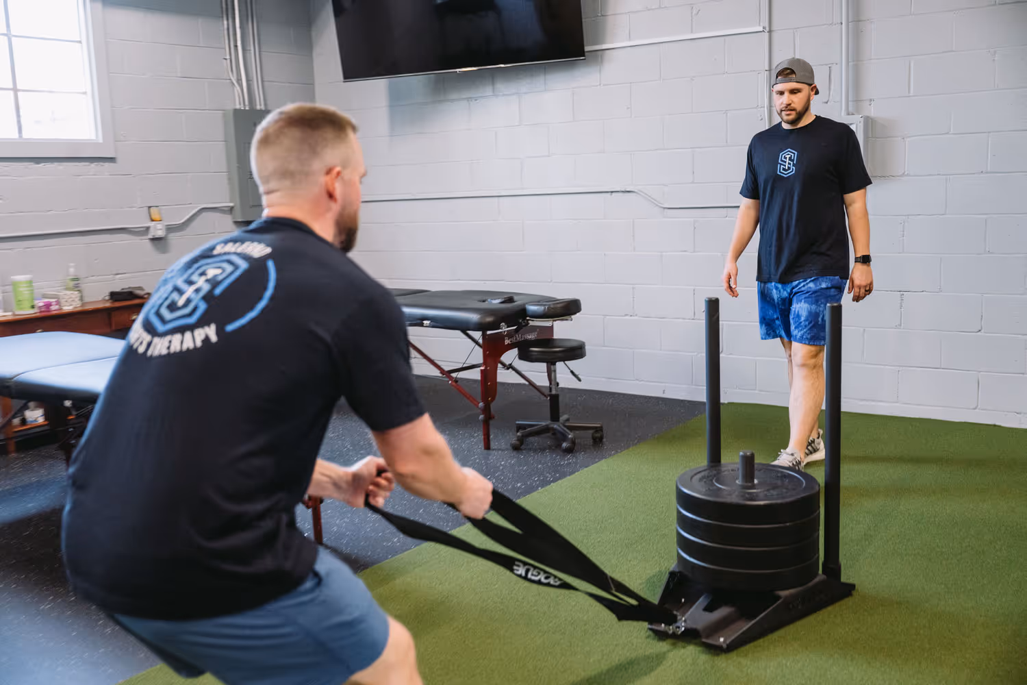 Two men in a gym; one is dragging a weighted sled while the other watches on artificial turf.