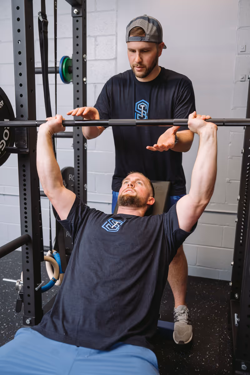 Man bench pressing a barbell with a spotter standing behind, both wearing black shirts and in a gym setting.