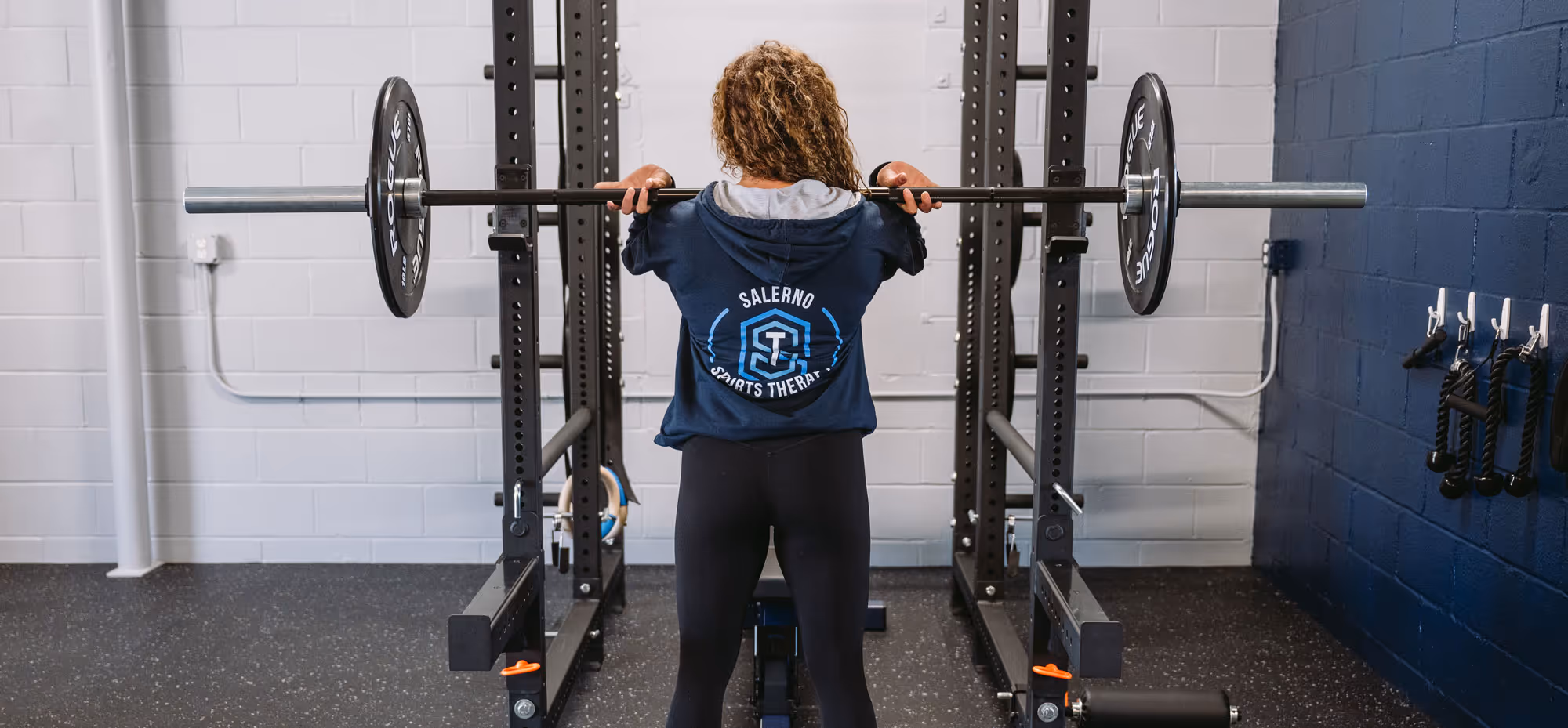 Person with curly hair lifting a barbell on their shoulders in a gym rack, wearing a blue hoodie with Salerno Sports Therapy logo on the back.