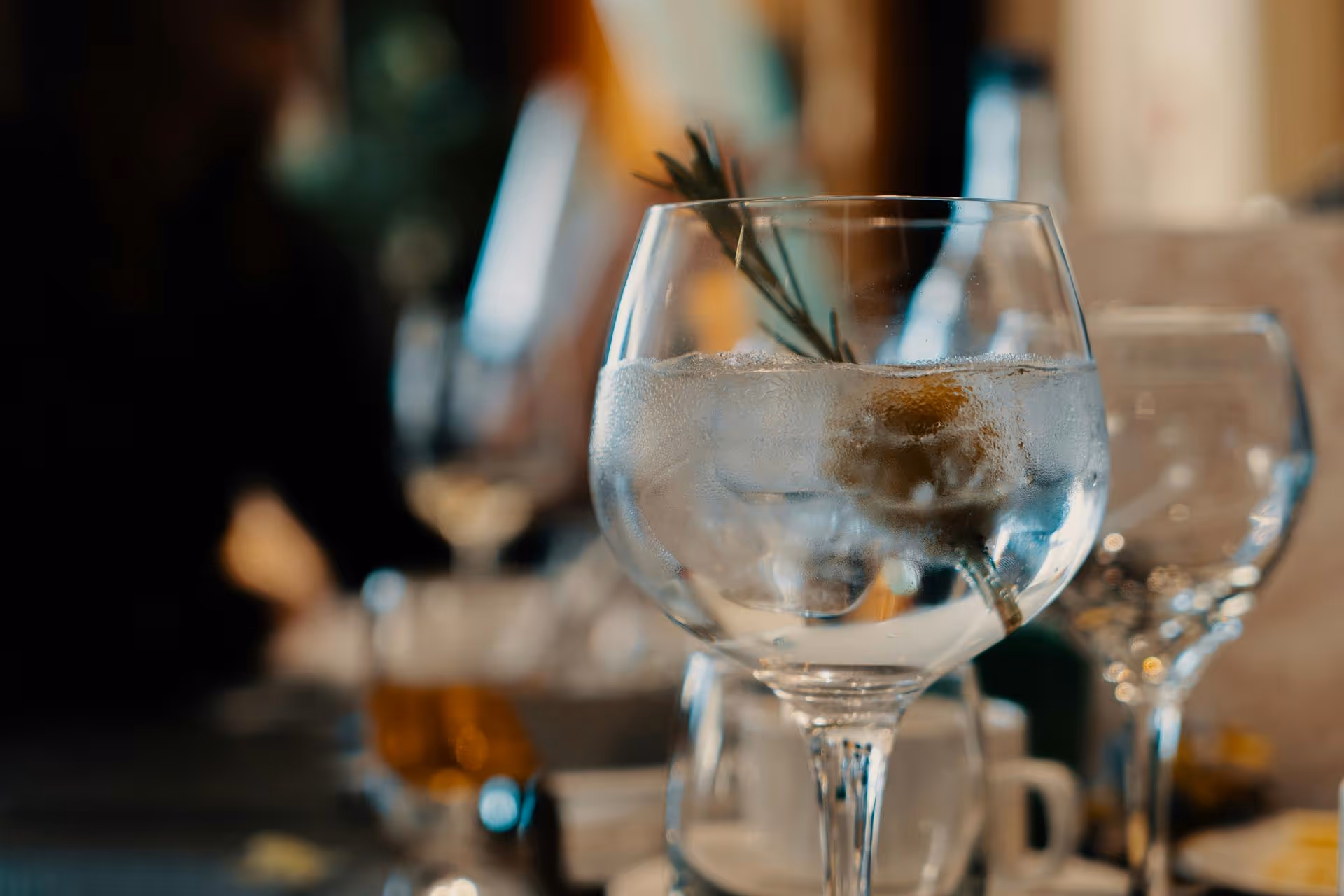 Close-up of a glass of clear liquid with a sprig of herbs, surrounded by blurred glasses on a table, creating a cozy dining atmosphere.