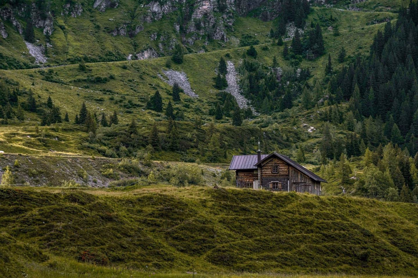 A quaint wooden cabin nestled on a green hillside, surrounded by lush mountains and a winding path in a serene landscape.