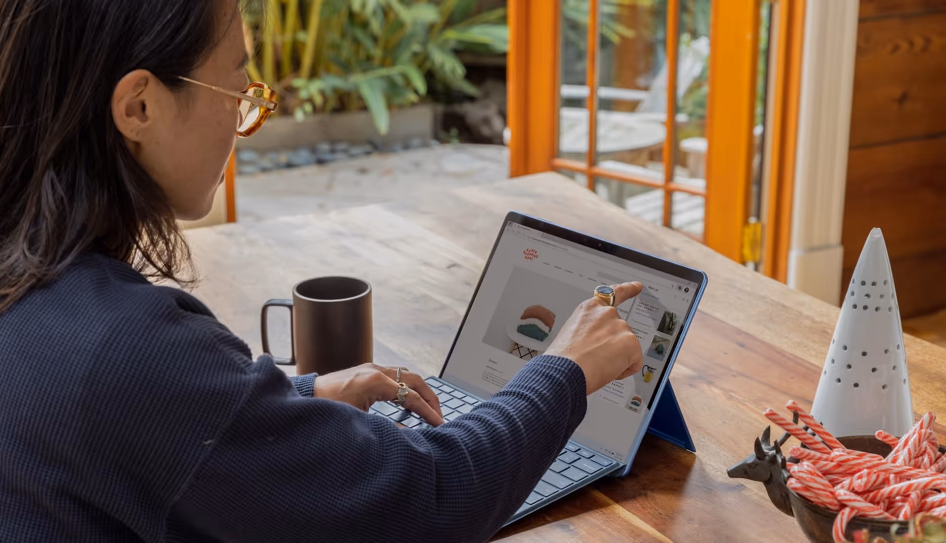 Person wearing glasses and a dark sweater using a tablet with keyboard on a wooden table near an open door.