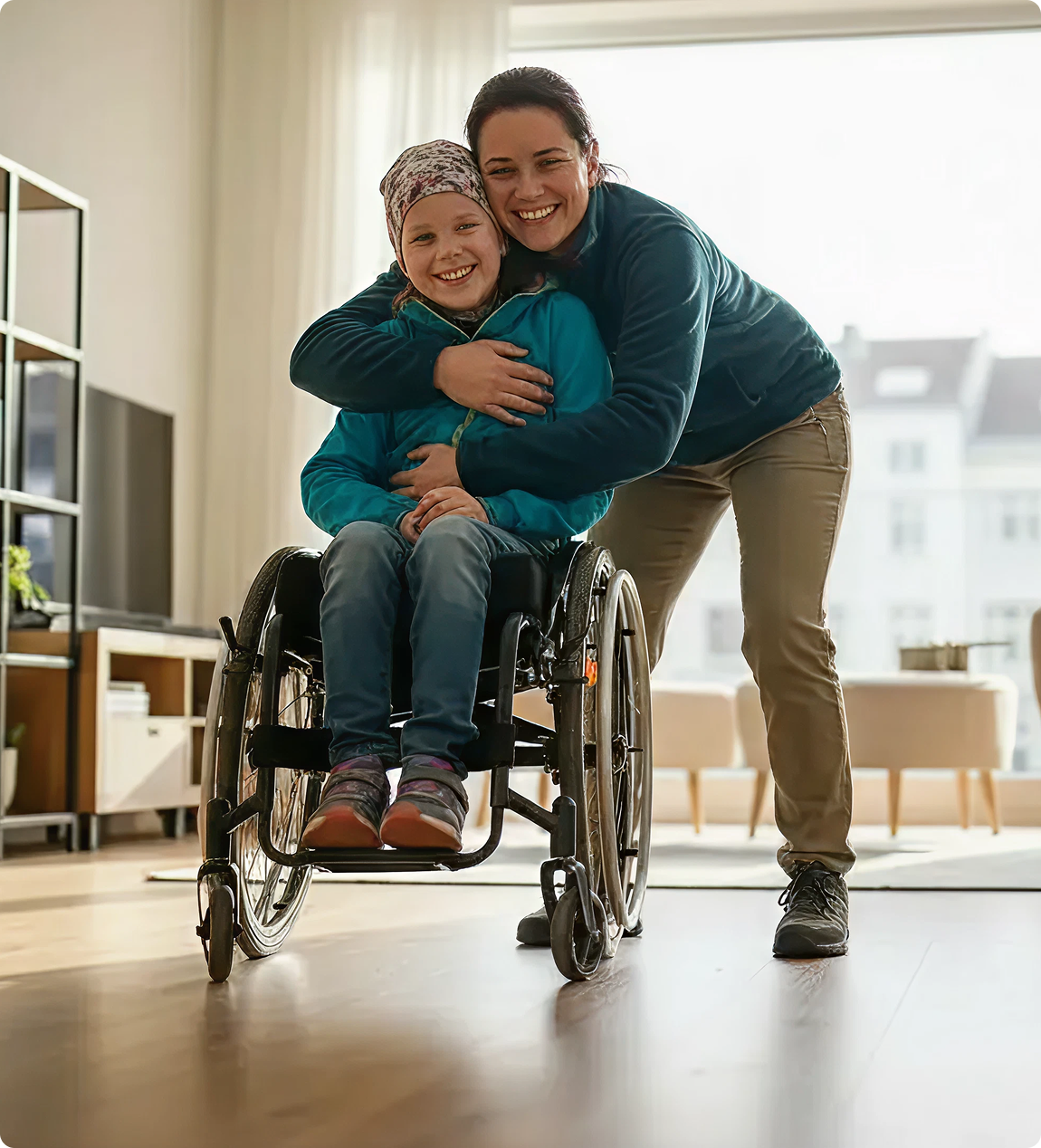 Woman in blue sweater smiling and hugging a child in a wheelchair wearing a patterned beanie and turquoise jacket.