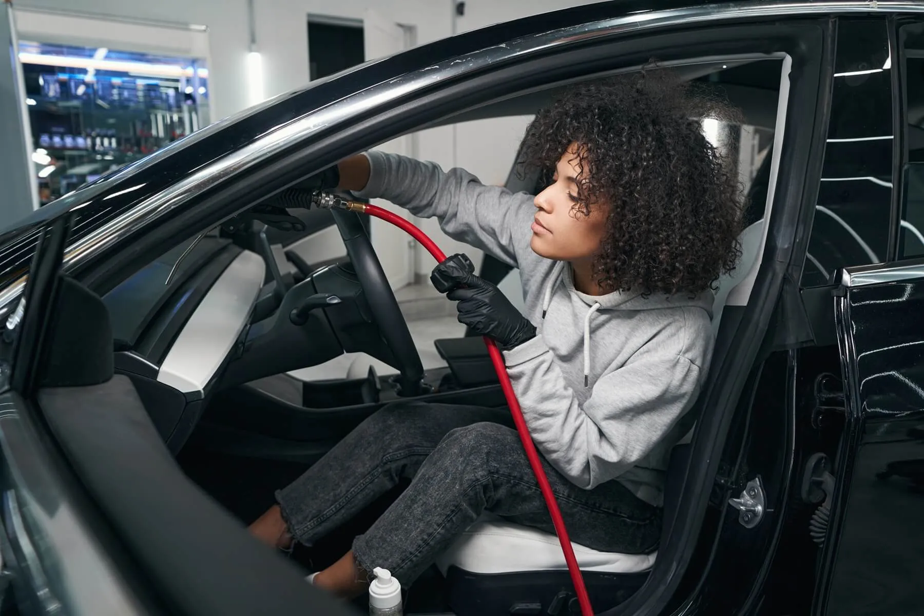 Woman wearing black gloves cleaning the interior windshield of a black car using a red hose tool.