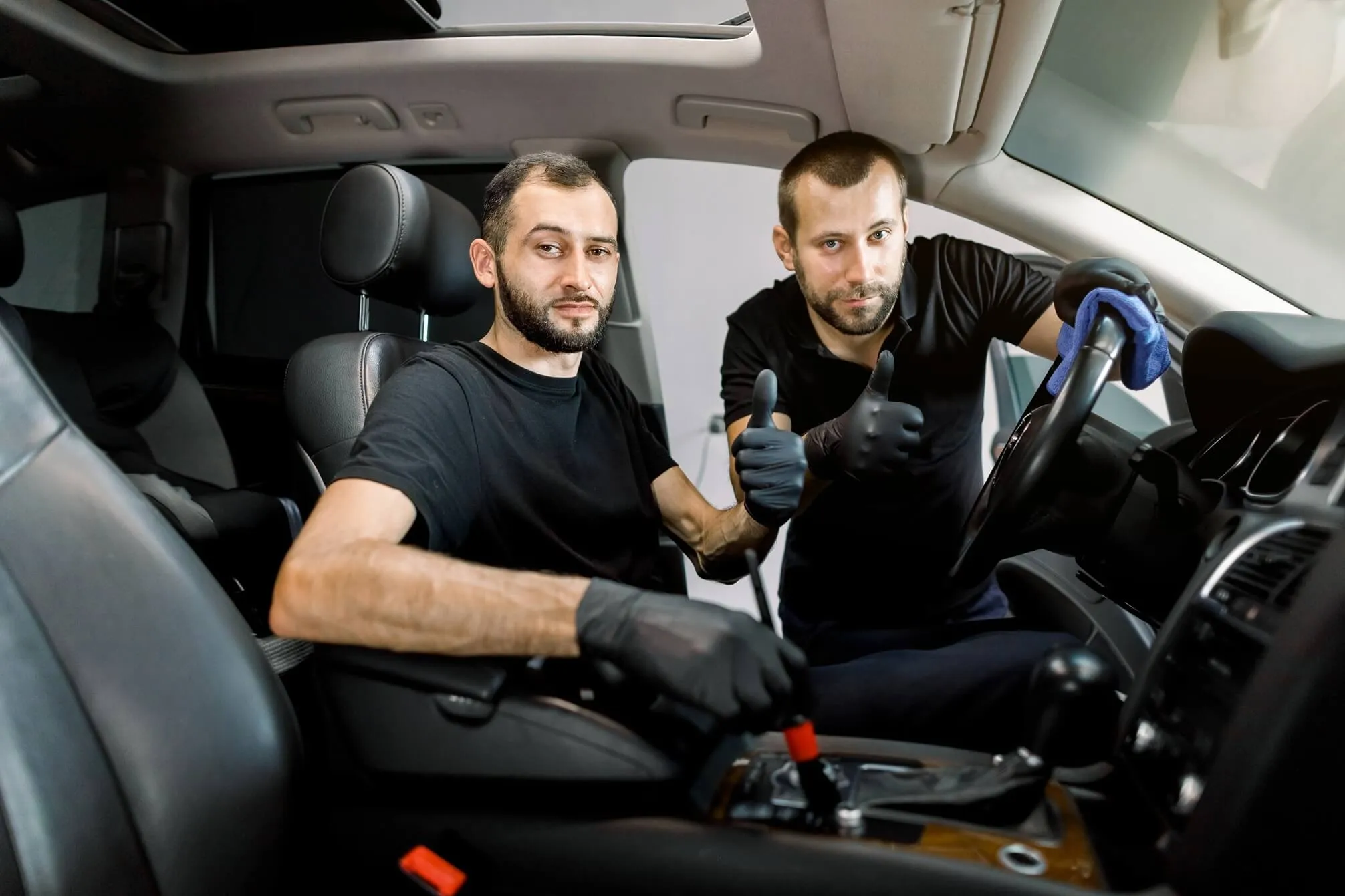 Two men wearing black gloves giving thumbs up gesture inside a car, one cleaning the steering wheel and the other shifting gears.