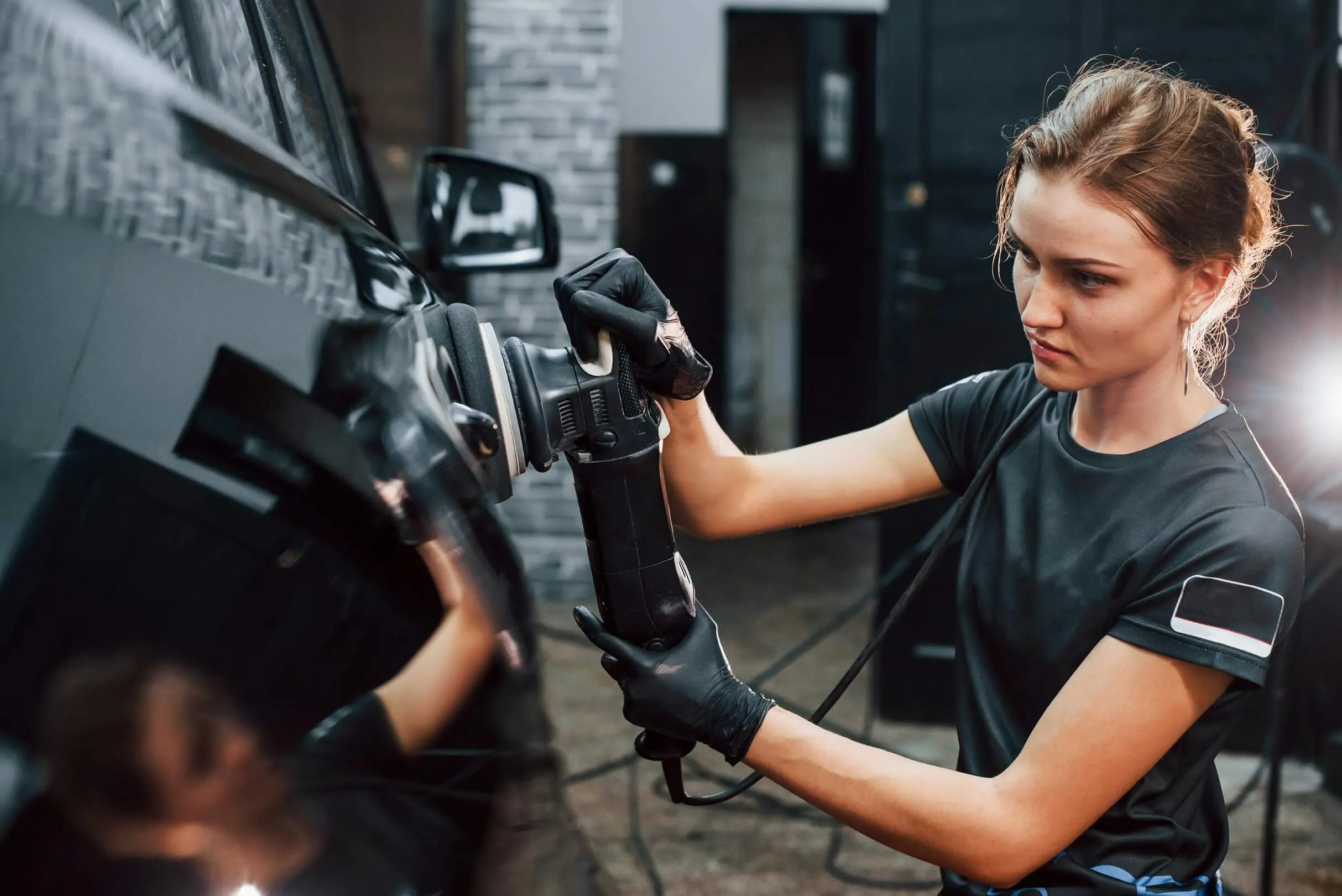 Woman wearing black gloves polishing a black car using a power buffer in a garage.