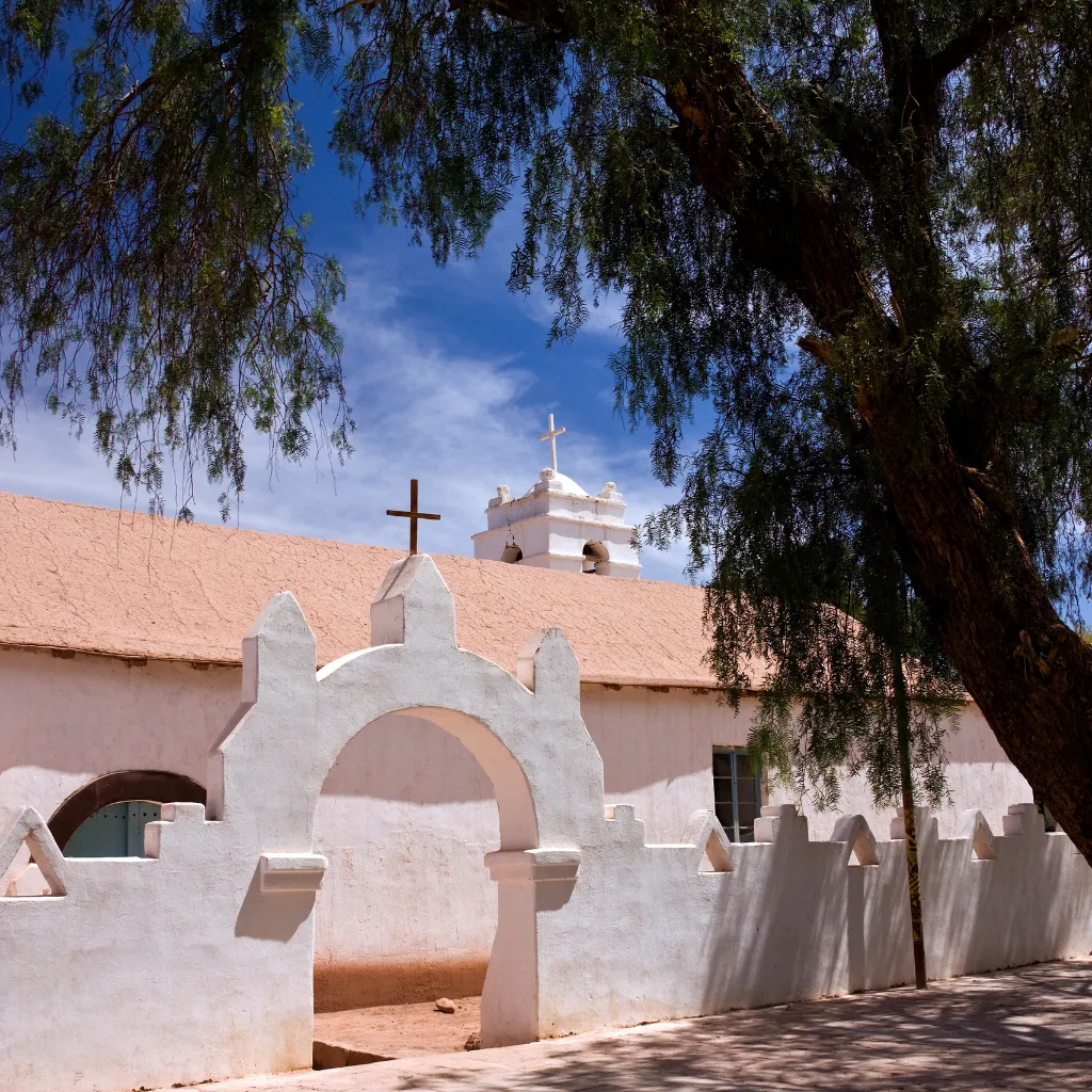 Fachada blanca de una iglesia antigua con cruces y un árbol grande a la derecha bajo un cielo azul  en San Pedro de Atacama