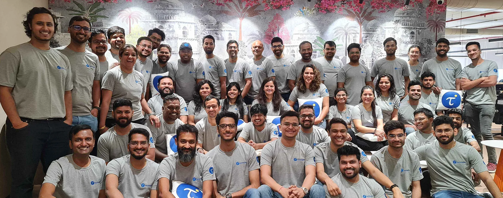 Large group of smiling people wearing matching gray t-shirts with a blue logo seated and standing in front of a decorative wall.