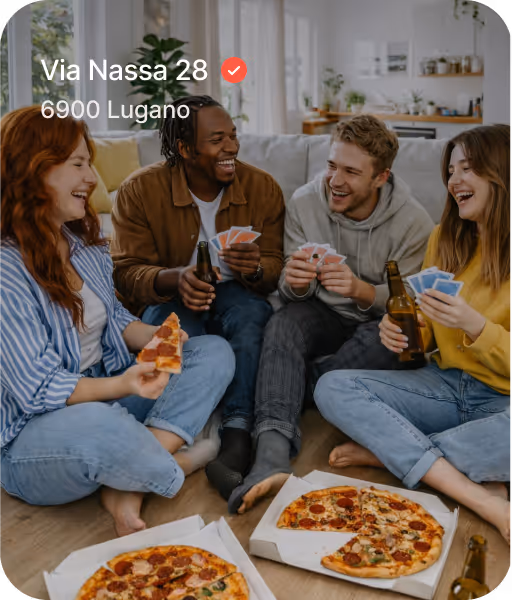 Four friends sitting on the floor playing cards and eating pizza while holding beer bottles in a cozy living room.