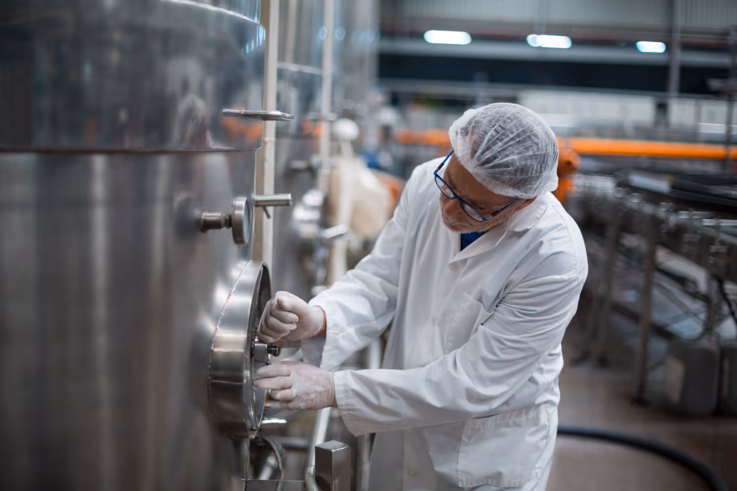 Worker in white lab coat, hairnet, gloves, and glasses operating stainless steel industrial equipment.
