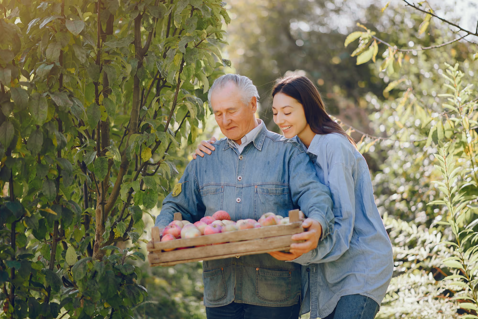 An elderly man holding a wooden crate full of apples with a young woman smiling beside him among green trees.