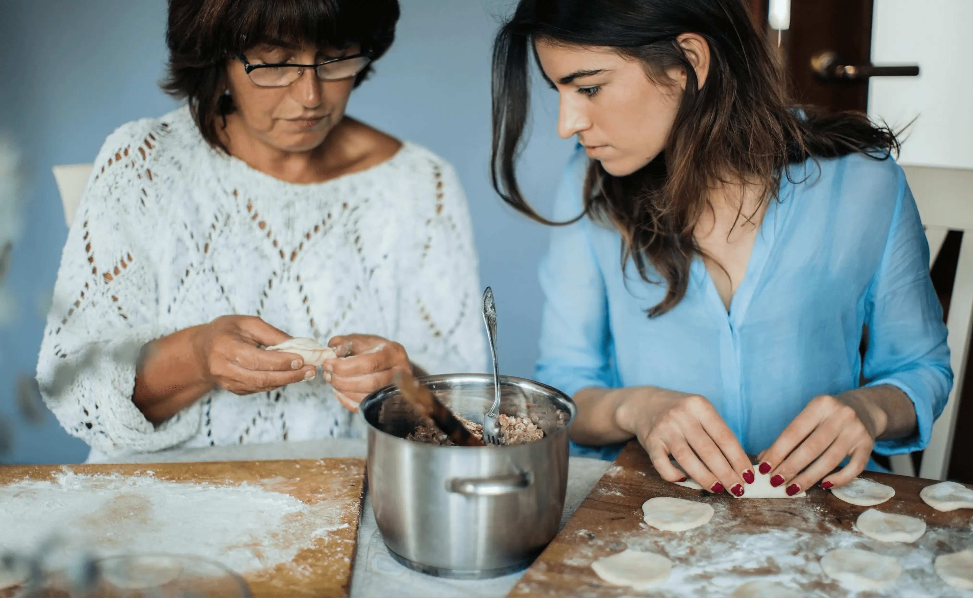 4: A middle-aged woman and a young woman are making dumplings together in a kitchen. The older woman is focused on shaping the dough, while the younger woman is concentrating on the filling.