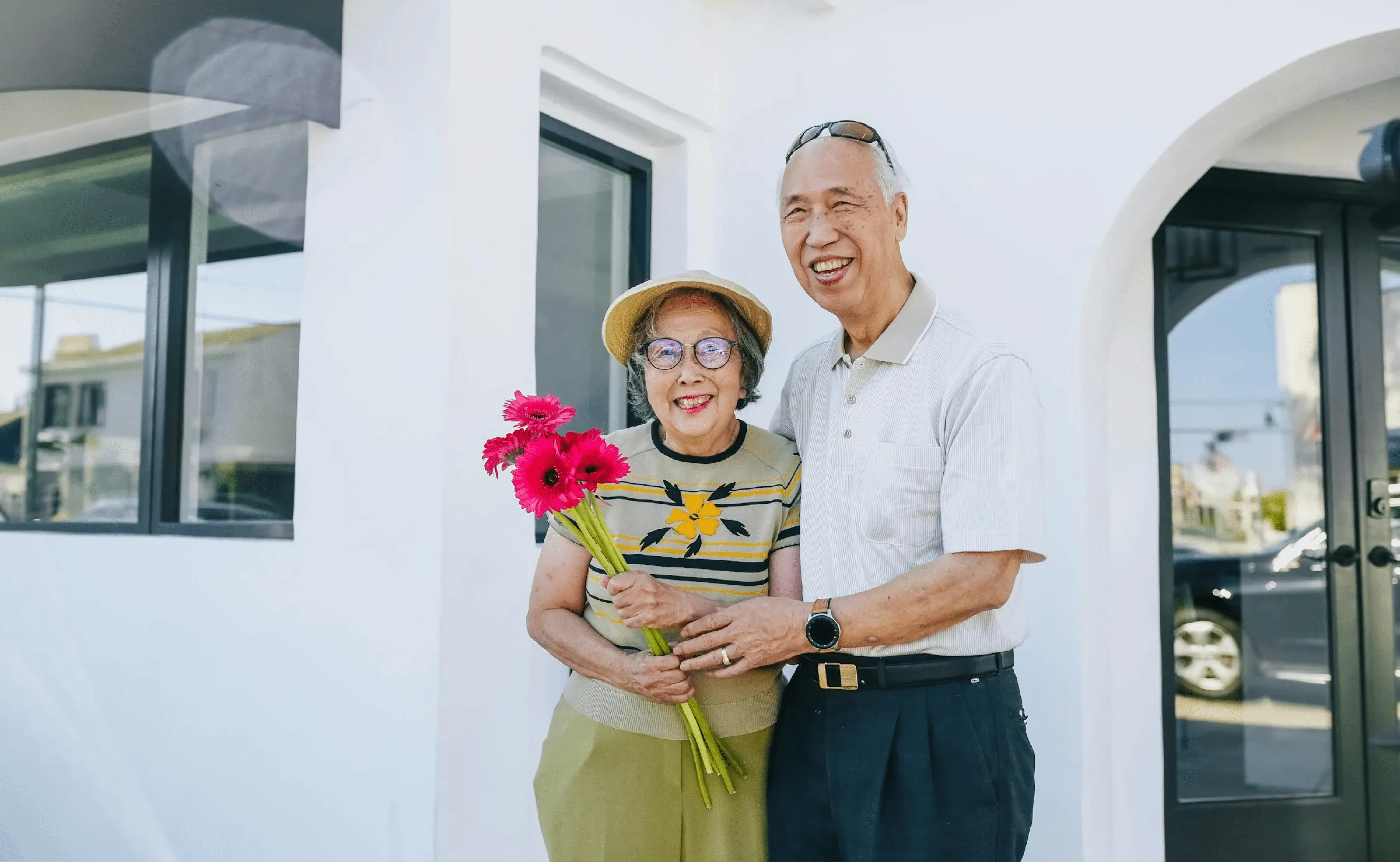 An elderly couple stands outside a modern white house. The woman is holding a bouquet of bright red flowers, and both are smiling joyfully at the camera.