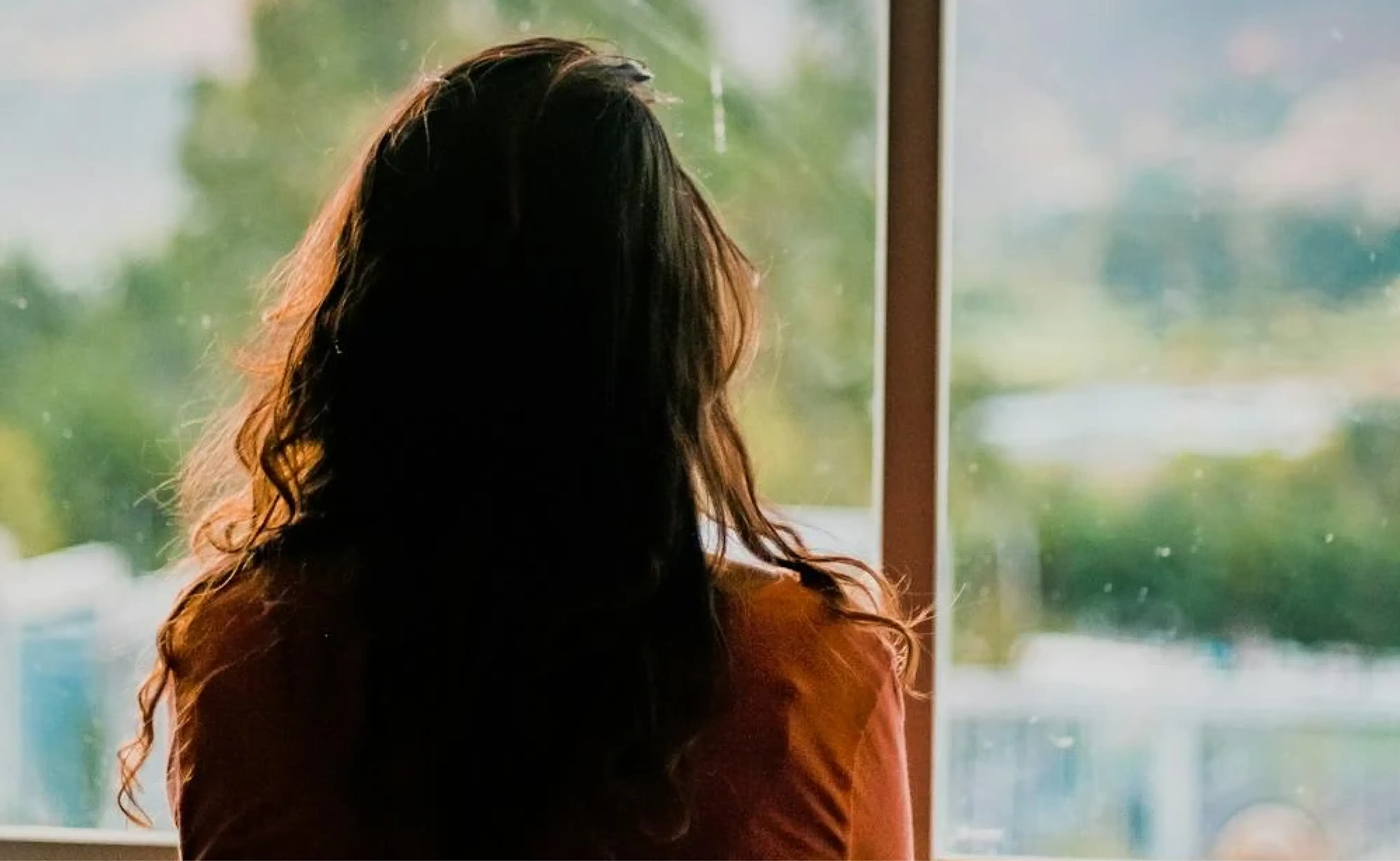 A woman with long hair looks thoughtfully out of a window. The background shows a blurred view of greenery, indicating she might be reflecting.