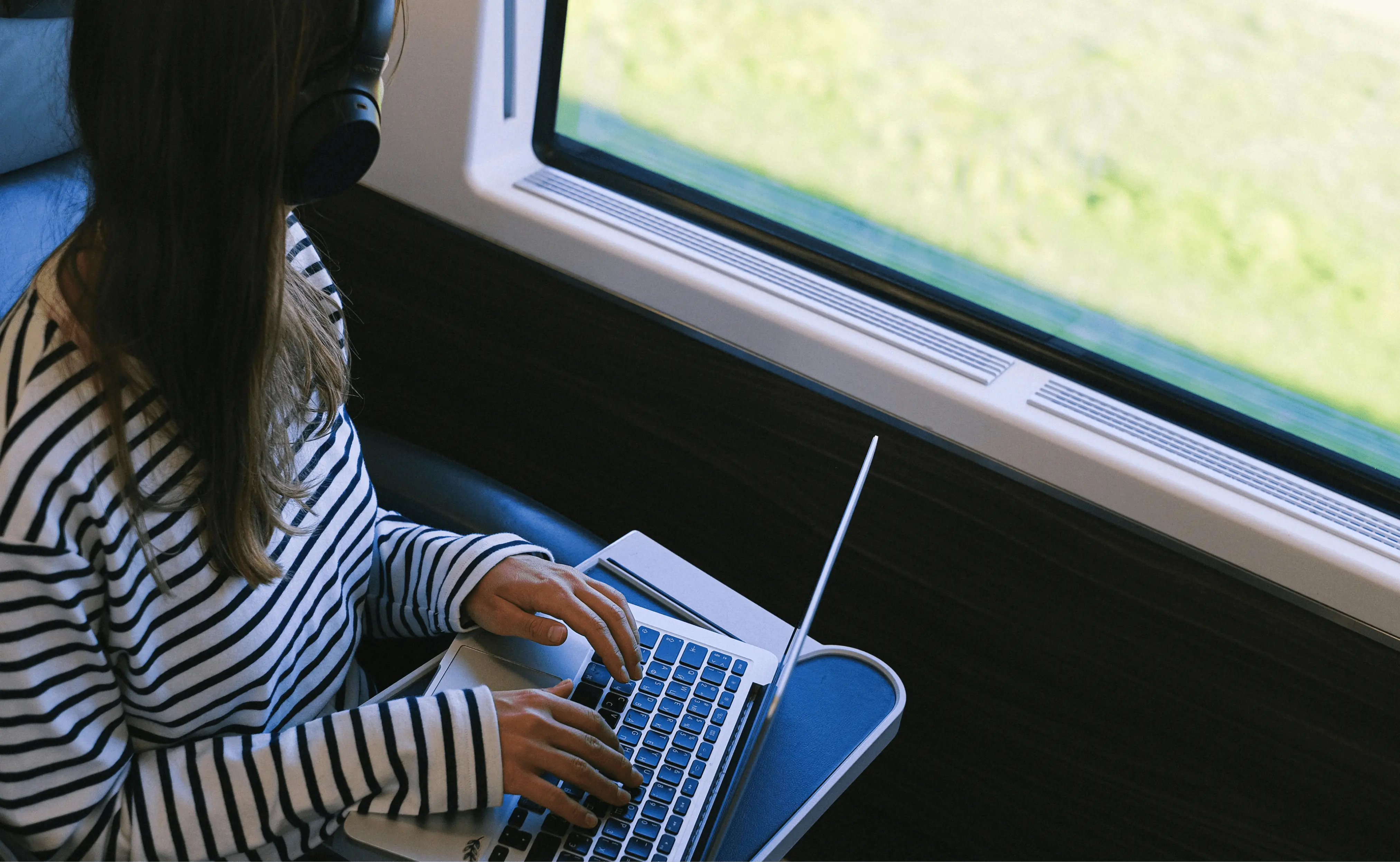 A person wearing a striped shirt and headphones is working on a laptop while seated on a train.