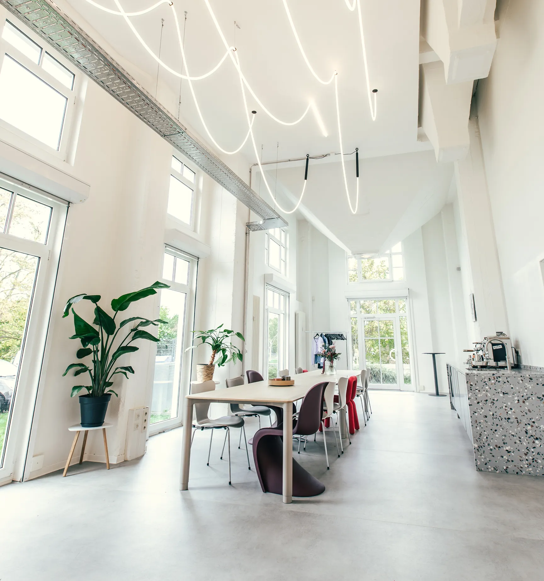 Helles modernes Büro mit großem Besprechungstisch, bunten Stühlen, großen Fenstern, Pflanzen und einer Kaffeemaschine auf der rechten Seite.