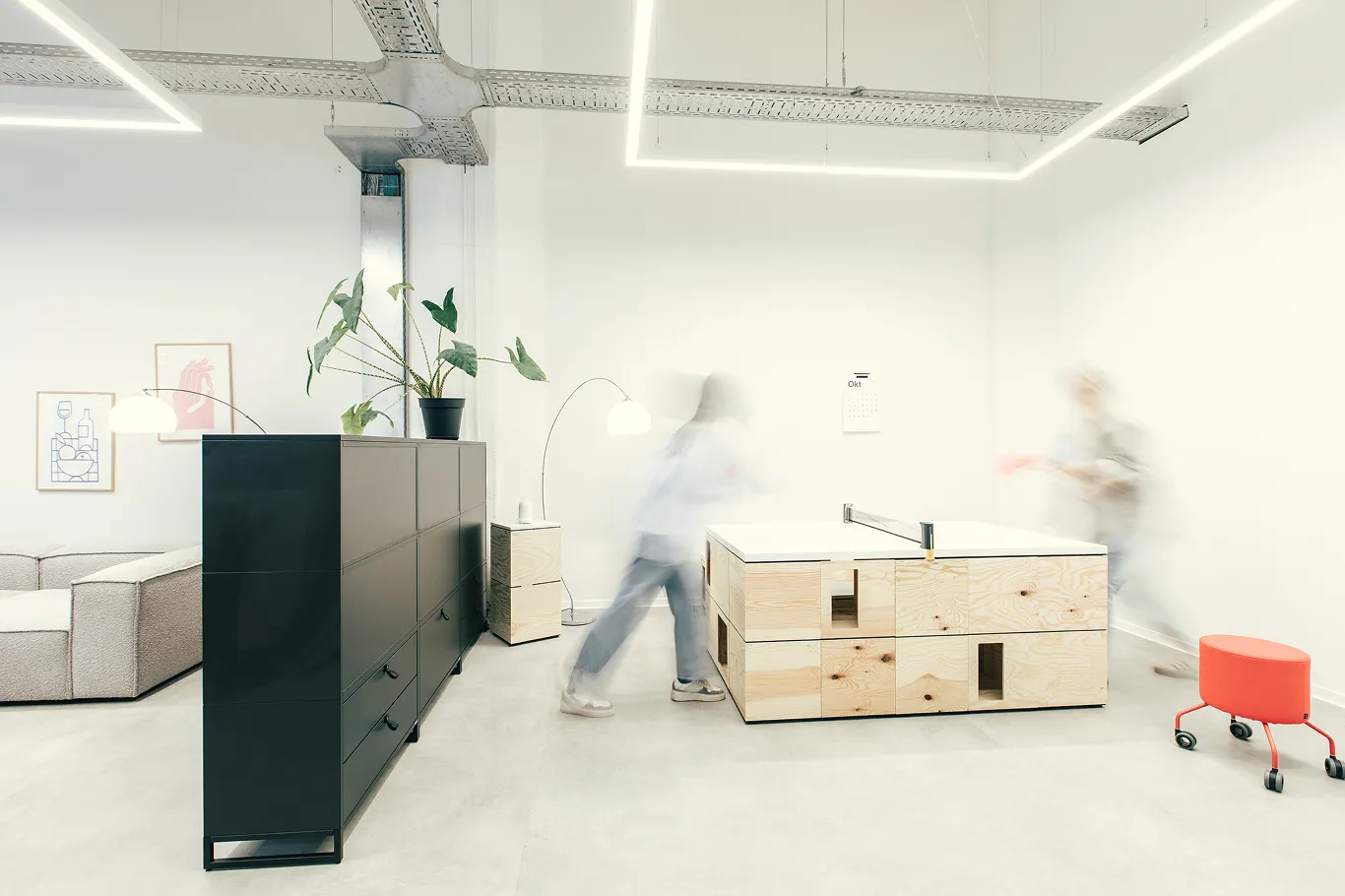 Moderner Bürobereich mit grauer Couch, schwarzem Sideboard, großem Holztisch und zwei Personen, die Tischtennis spielen.