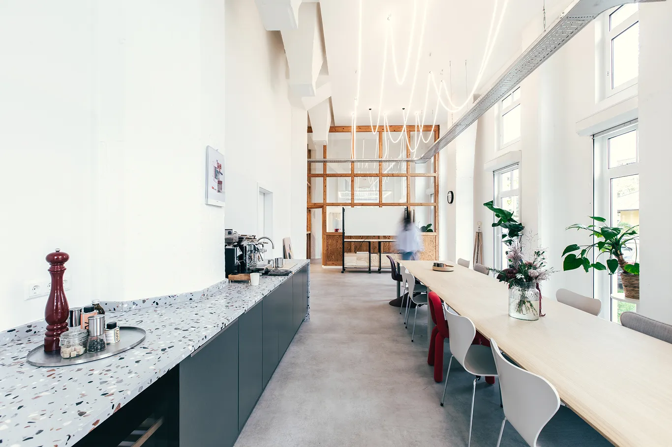 Heller, moderner Konferenzraum mit langem Holztisch, bunten Stühlen, Pflanzen am Fenster und einer Kaffeemaschine auf der linken Theke.