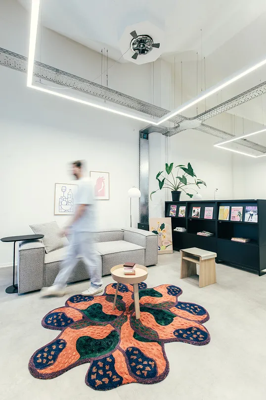 Moderner, heller Wohnraum mit grauem Sofa, buntem Teppich in organischer Form, niedrigen Holztischen und schwarzem Regal mit Pflanzen und Büchern.