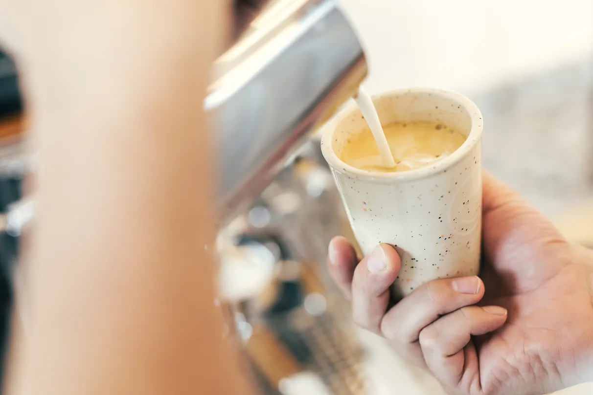 Hand hält eine Tasse, während Milch in Espresso gegossen wird, um einen Kaffee zuzubereiten.