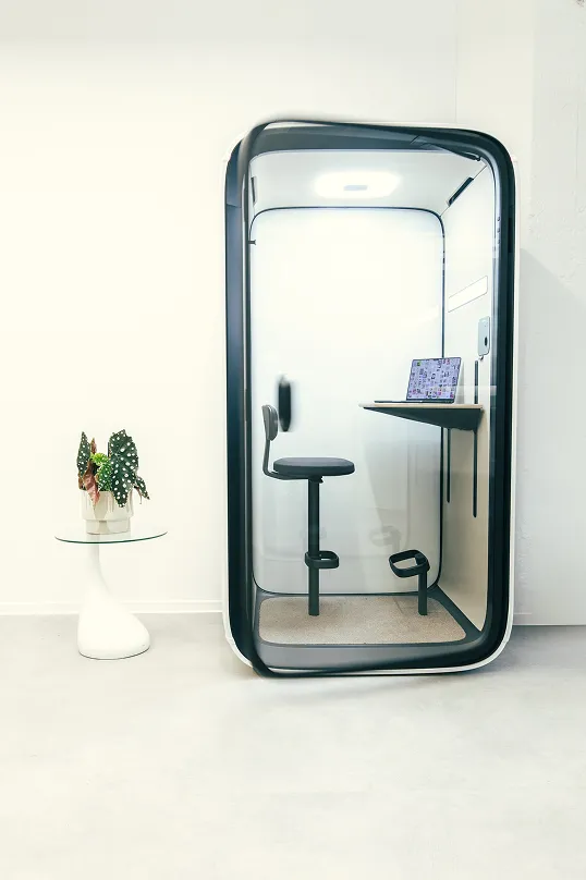 Moderner einzelner Telefon- oder Arbeitsraum mit schwarzem Drehstuhl, Holztisch und Laptop neben einem kleinen weißen Glasbeistelltisch mit einer Topfpflanze.