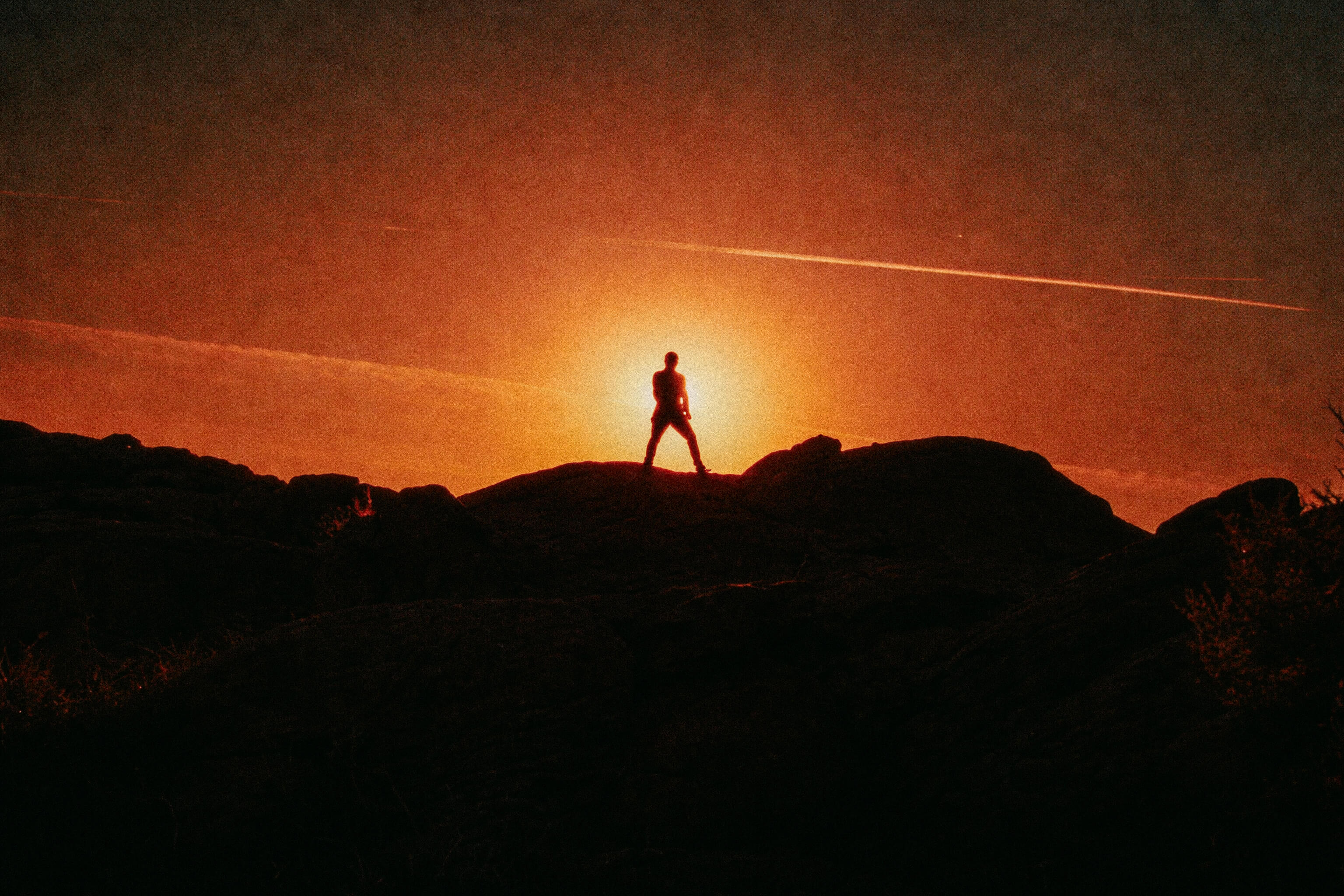 Silhouette of a person standing on rocks with the sun setting behind them, creating an orange and red sky.