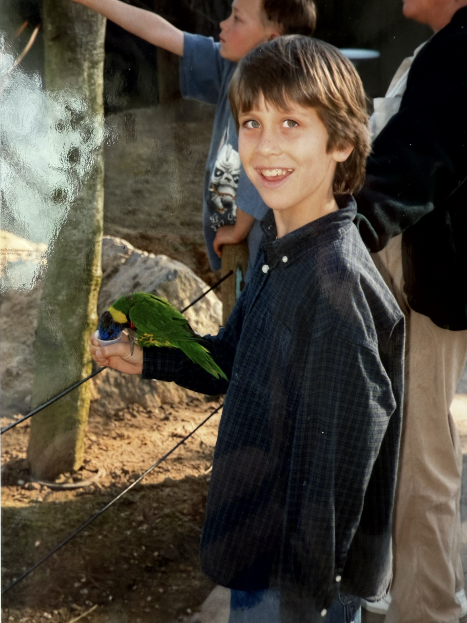 Smiling boy in a dark checkered shirt holding a green parrot while looking at the camera at an outdoor setting.