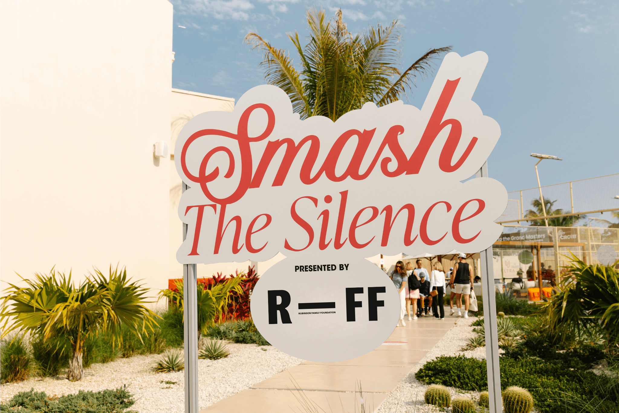 Outdoor sign reading 'Smash The Silence presented by RFF Robinson Family Foundation' with people walking on a path and palm trees in the background.