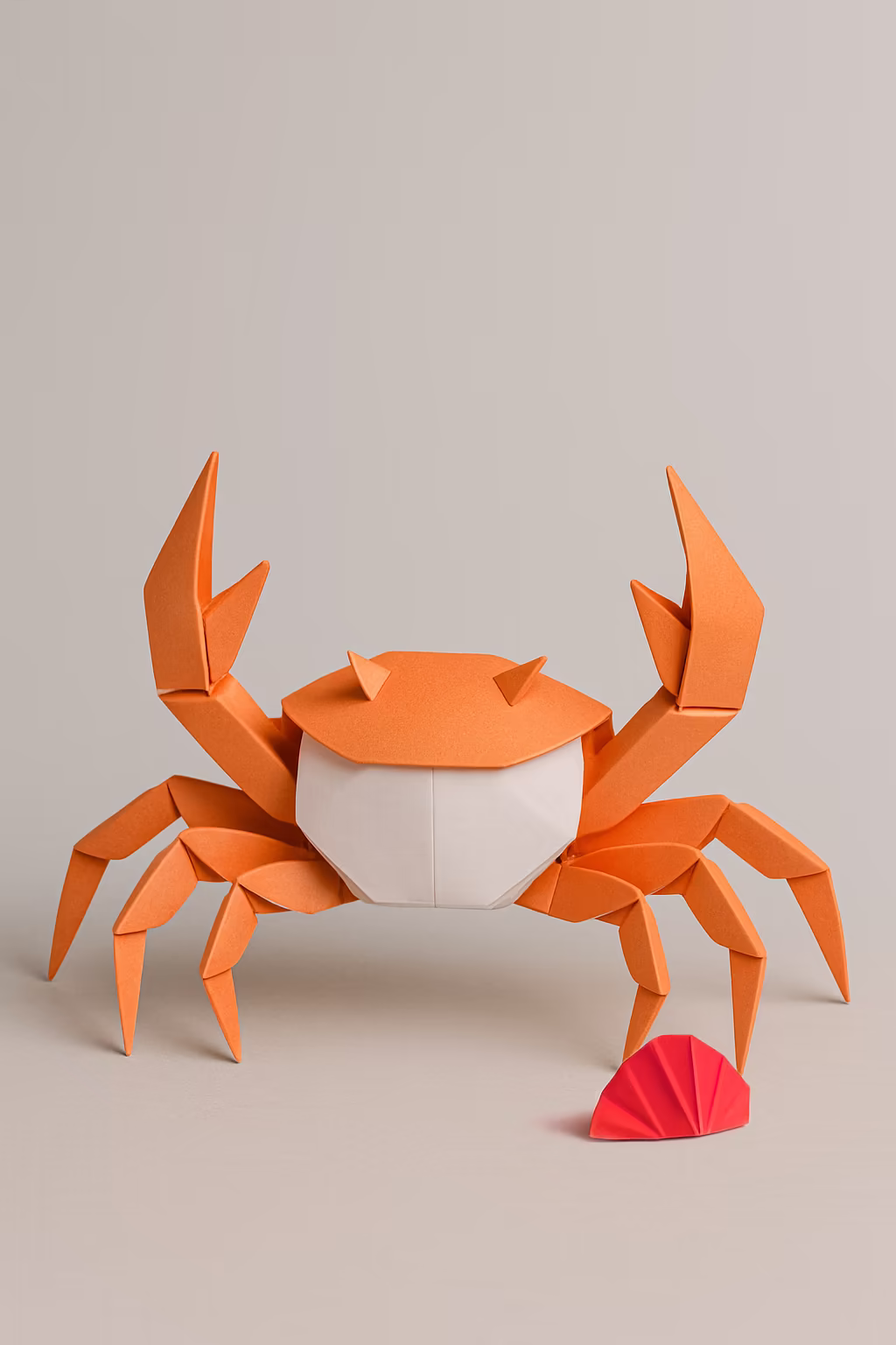 Orange and white origami crab with raised claws and a small red origami shell beside it on a neutral background.