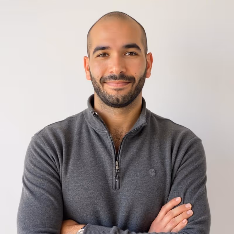 Smiling bald man with beard wearing a grey zip-up sweater, standing with arms crossed against a white background.
