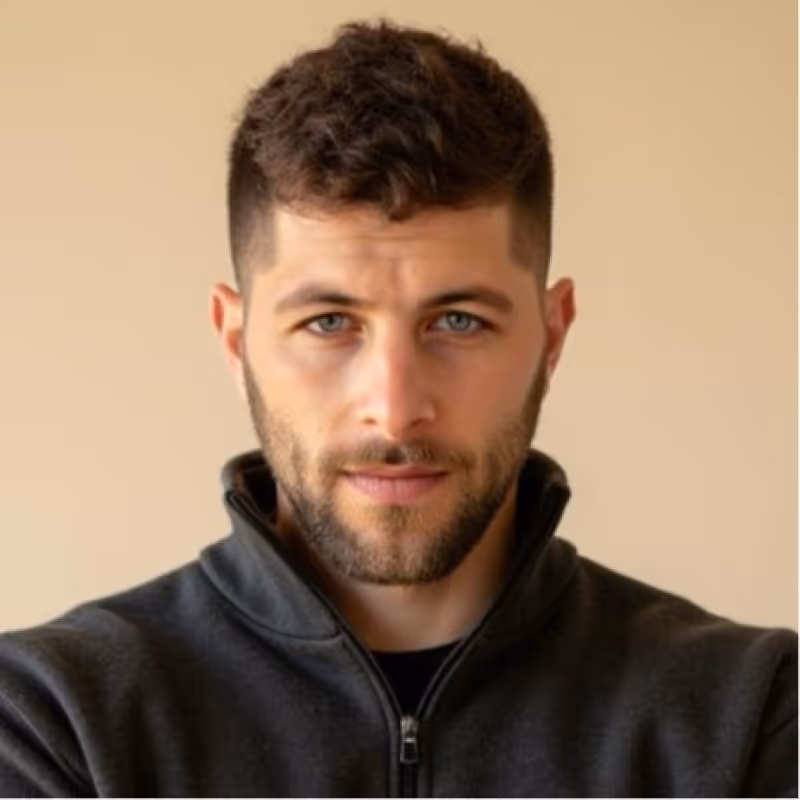 Close-up portrait of a man with short curly brown hair, blue eyes, and a beard wearing a dark zip-up jacket against a beige background.