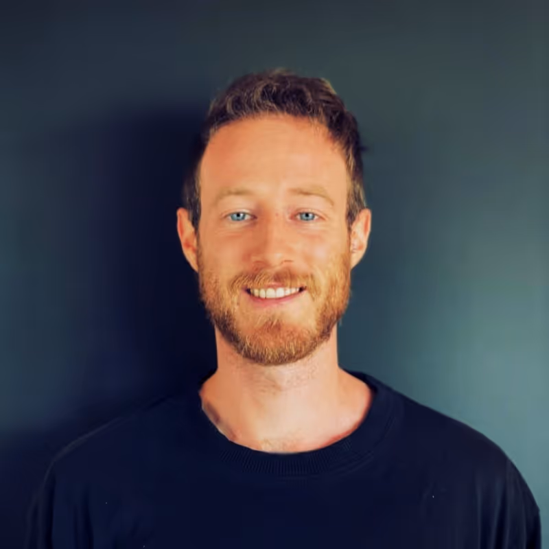 Smiling man with light brown curly hair and beard wearing a black shirt against a dark background.