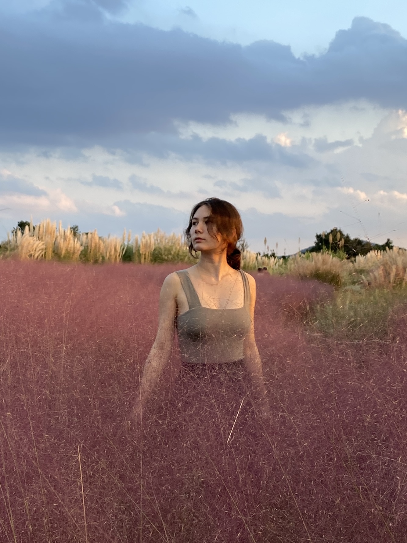 Young woman in a sleeveless top standing in a field of tall pink grass under a cloudy sky at sunset.