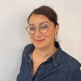 Portrait d'une femme souriante portant des lunettes rondes et une chemise en jean foncé, avec les cheveux attachés en arrière.