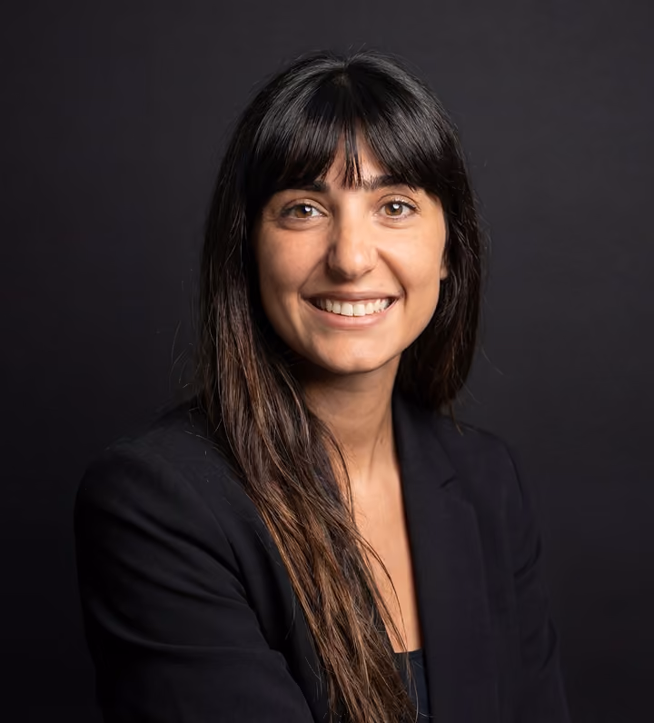 Portrait d'une femme souriante aux longs cheveux bruns, portant un blazer noir devant un fond sombre.