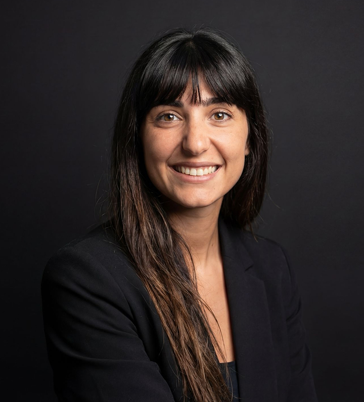 Portrait d'une femme souriante aux longs cheveux bruns, portant un blazer noir devant un fond sombre.