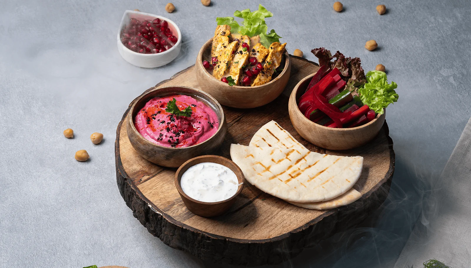 Wooden board with pita bread, pink hummus, yogurt dip, grilled chicken, and vegetable sticks in bowls.