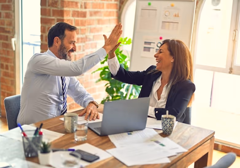 Two business colleagues smiling and high-fiving each other at a desk with laptop and papers.