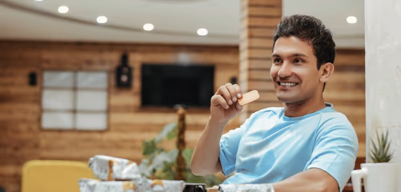 Smiling man in blue shirt holding a snack in a cozy, modern room with wooden walls.