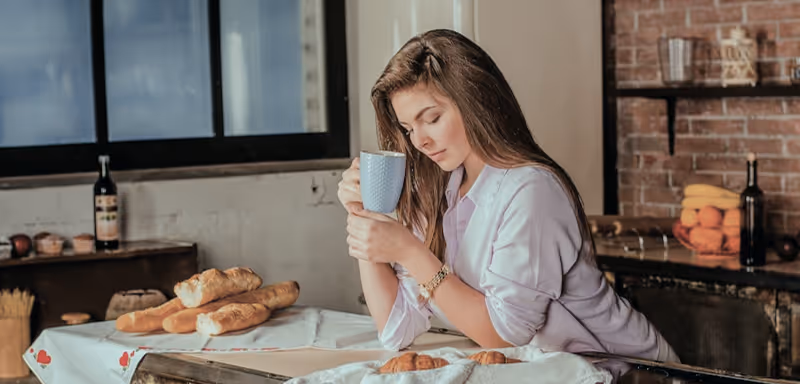 Woman holding mug sits at table with croissants and baguettes in cozy kitchen setting.