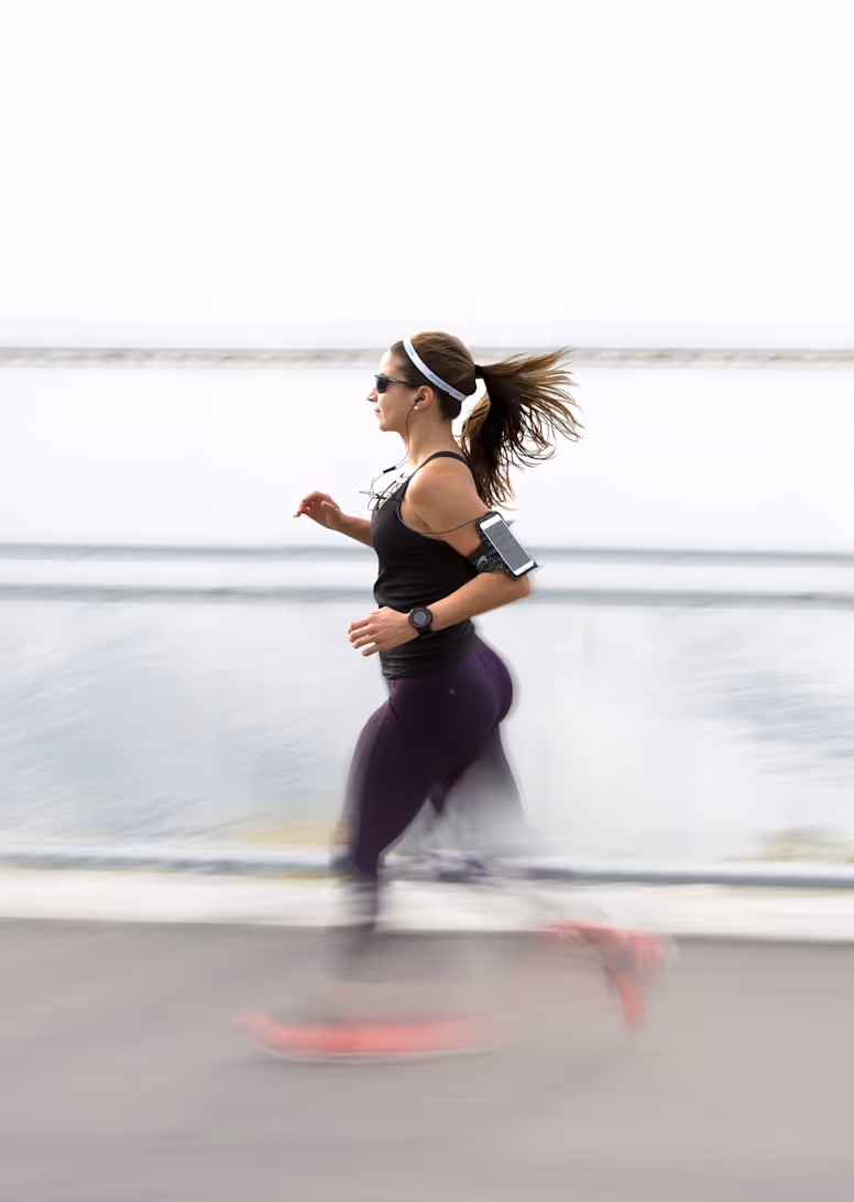 Woman with ponytail running outdoors near water wearing sunglasses and wireless earphones.