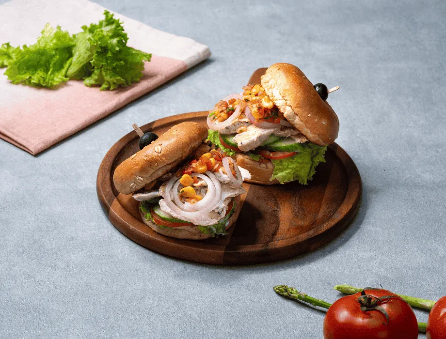 Two chicken sandwiches with lettuce, tomato, onion, and corn on a wooden plate with a pink napkin.
