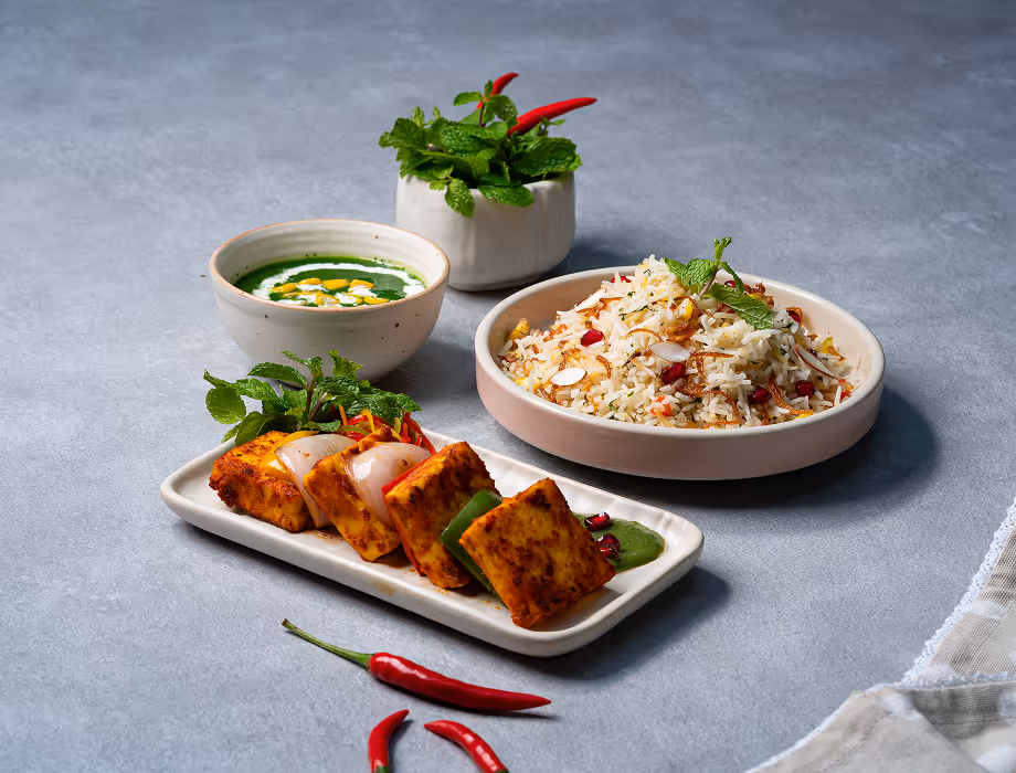 Plate of grilled paneer cubes with vegetables, bowl of rice with pomegranate, and green chutney with cream.