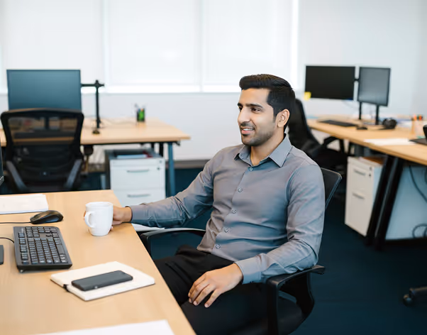 Man in gray shirt sitting at office desk holding a white coffee mug and smiling.