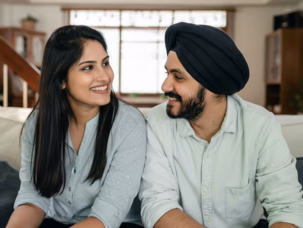 Smiling man with turban and woman with long hair sitting on a couch, looking at each other.