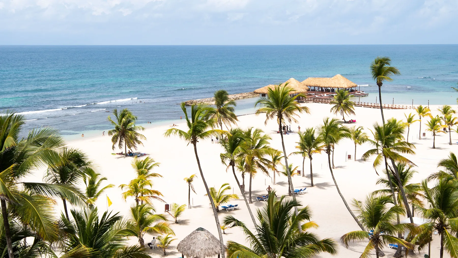 Palm trees on a white-sand beach beside turquoise water in Juan Dolio, with thatched sun umbrellas
