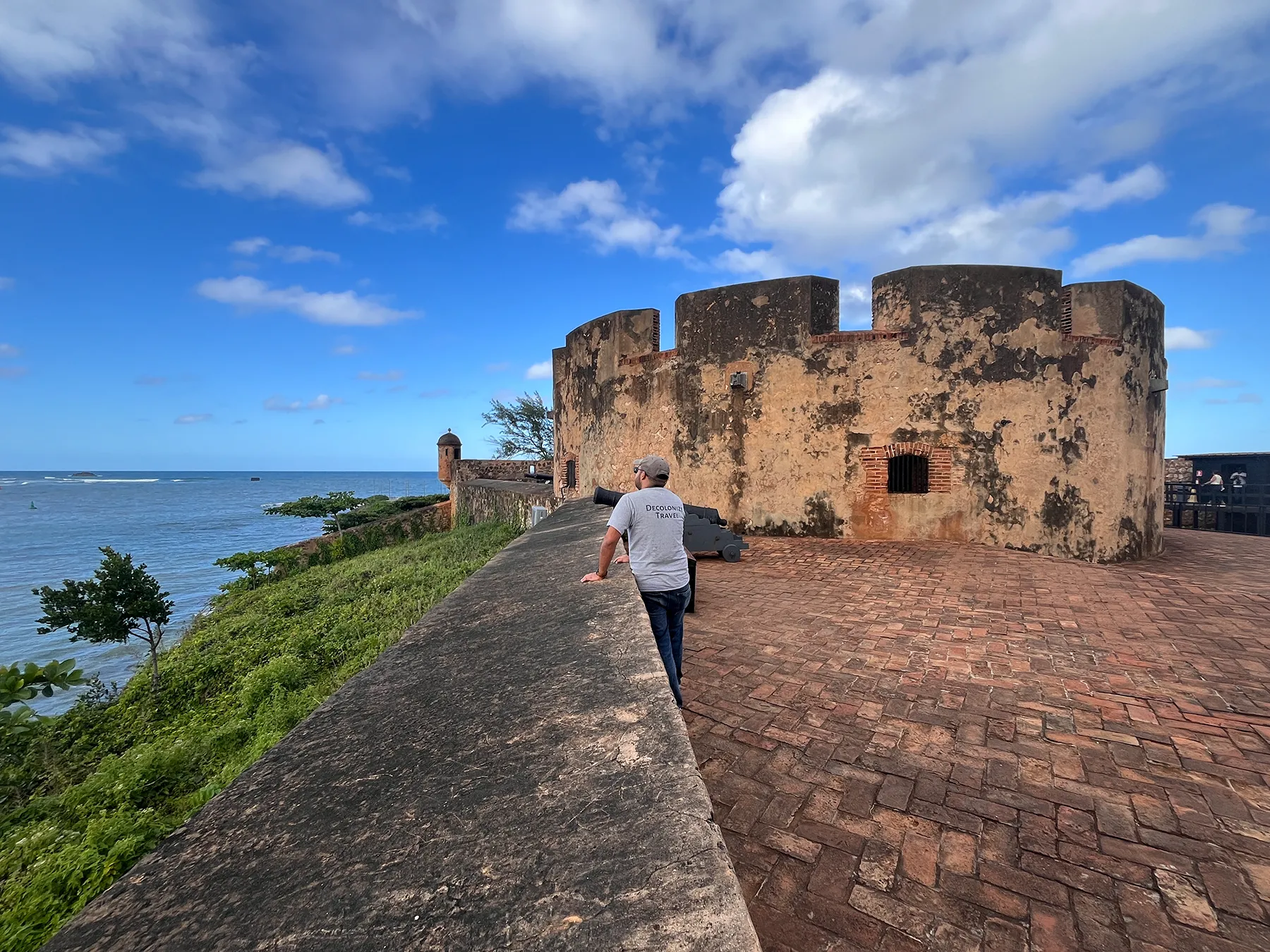 A person walking toward a historic stone fort tower on a grassy coastal point in the Dominican Republic
