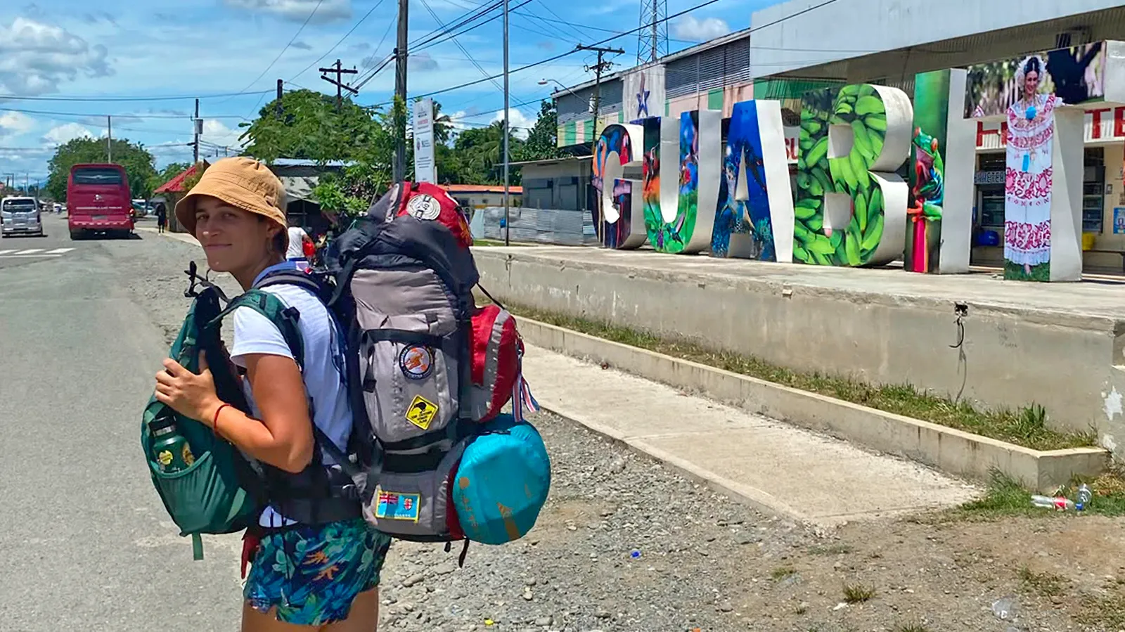 Daniela carries a large hiking backpack on a Costa Rica roadside, beside a colorful town sign